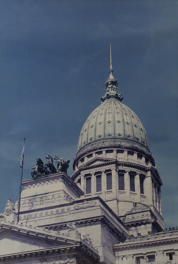  La fotografía presenta en primer plano, un acercamiento a la gran cúpula del Congreso de la Nación. La cúpula ocupa el centro de la composición y está decorada con relieves circulares y detalles ornamentales que resaltan sobre el cielo azul profundo del fondo. En la parte superior se eleva una aguja fina que acentúa la verticalidad de la estructura. Debajo de la cúpula se distinguen columnas y cornisas clásicas, mientras que en el lateral izquierdo aparece una bandera argentina ondeando suavemente. Sobre una de las esquinas del edificio se observan esculturas ecuestres oscuras, que aportan dramatismo a la escena. La fotografía utiliza una perspectiva cerrada que concentra la atención en los detalles arquitectónicos y transmite una sensación de majestuosidad.